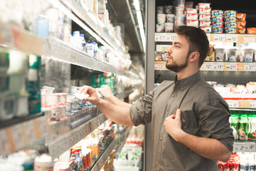 Happy man wearing a shirt stands at the refrigerator in the milk department of the supermarket and takes yogurt from the shelf. Buyer with a beard takes a yogurt from his fridge at a grocery store.