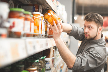 Adult man takes a can of vegetables with a shelf from a grocery store. Choosing and buying canned...