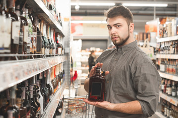 Portrait of a man with a beard standing in a supermarket with a bottle of a cognac in hands and looking at the camera.Buyer man posing at the camera with strong drink bottle in the supermarket aisle