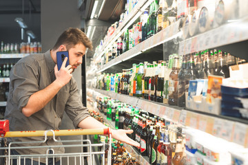 Business man stands with a cart in a supermarket, chooses a beer bottle on the shelves in the alcohol department and calls the phone.Man asks a friend by phone to take a beer in the store.Beer choice