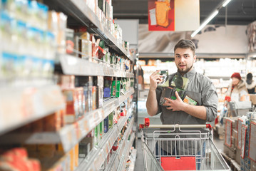 Happy man stands with a cart in the aisle of a supermarket, holds in his hands many packages at a...