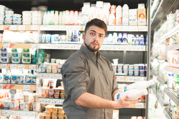 Surprised man with a beard is in the milk department of a supermarket with bubbles of milk in his hands,looking at the camera in amazement.Portrait of an amusing buyer buys products at a grocery store