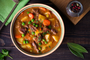 Homemade Irish beef stew with potatoes, carrots and peas in bowl on wooden table. overhead,...