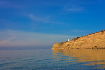 Fototapeta na wymiar White small lighthouse on cliff by blue Greek sea