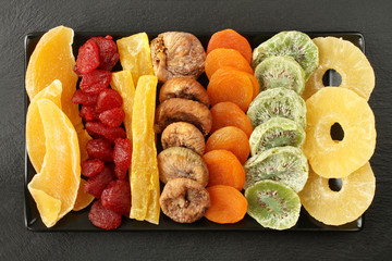 Candied fruit pieces of mango, papaya, kiwi, pineapple, dried strawberries, figs, apricots, lying on a black ceramic plate on a black stone surface. Macro