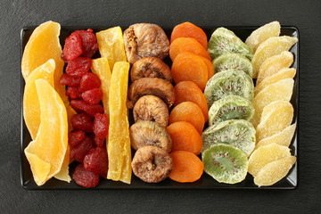Dried fruit pieces of mango, papaya, kiwi; dried strawberries, figs, apricots; pieces of candied ginger root, lying on a black ceramic plate on a black stone surface. Macro