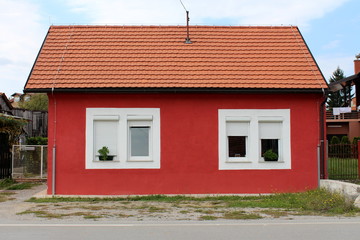 Completely renovated small suburban family house with new windows and roof tiles surrounded with uncut grass and gravel next to paved road on cloudy blue sky background