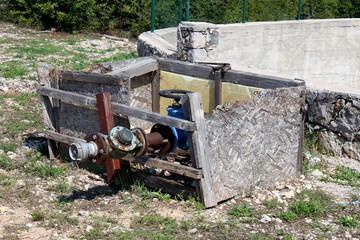 Fototapeta na wymiar Complex connection of multiple pipeline connectors with metal valve connected with strong screws next to large abandoned concrete water tank surrounded with grass and dense trees in background