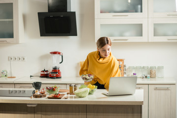 Fototapeta na wymiar Attentive woman using laptop in the kitchen while standing with salad