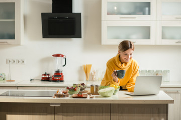 Happy woman with cup of tea using laptop in the kitchen