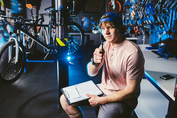 Young handsome stylish man in a cap snapback and with a tattoo small business owner selling a bicycle, the workshop sits on the background of the store in the hands of a notebook to write notes
