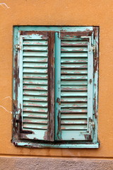 Dilapidated old closed wooden blinds with cracked blue paint mounted with rusted metal hinges on wooden frame surrounded with orange and purple facade of family house wall on warm sunny day