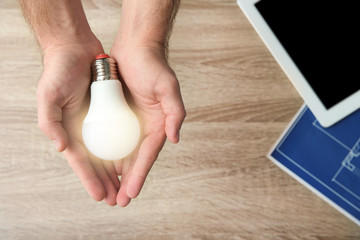 Man holding lamp bulb over table with tablet and blueprint, top view. Space for text