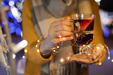 Woman with glass cup of mulled wine and garland at winter fair, closeup