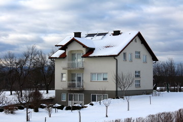 New suburban family house with two front balconies covered in snow surrounded with small and large trees on cloudy stormy day at sunset