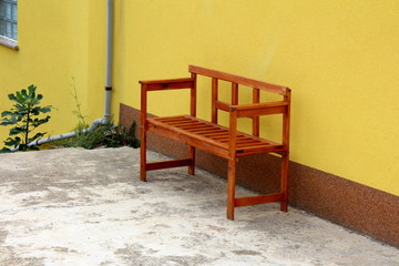 New wooden backyard bench left next to yellow house wall with plastic gutter pipe and plants on warm sunny day