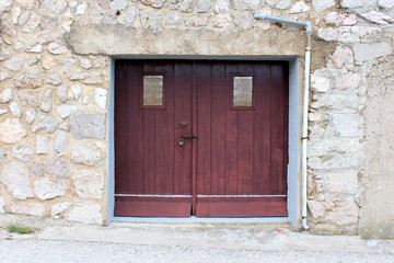 Old wooden garage doors made of wooden boards and rubber rain protection locked with rusted latch and padlock with two small windows mounted on traditional stone wall next to water pipe on warm cloudy