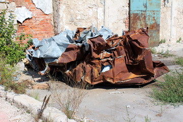 Pile of rusted crumpled sheet metal in front of abandoned cracked factory wall surrounded with rubble and grass on warm sunny day