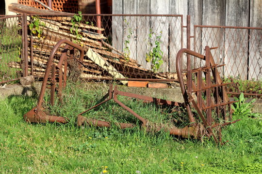 Rusted abandoned vintage agricultural farming equipment used to work with tractors on soil left in high grass in front of metal rusted fence and old barn on warm sunny day