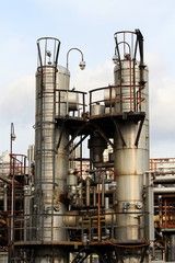 Rusted metal storage silos connected with complex pipeline network at abandoned industrial complex with cloudy blue sky background