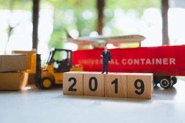 Miniature people, businessman standing on wooden block 2019 with logistic vehicle background using as business and logistics concept