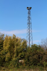Tall metal structure holding four large public civil defence warning air sirens above small completely overgrown structure and dense forest on clear blue sky background