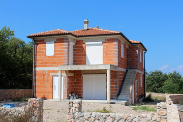 Unfinished red bricks family house with new doors and windows surrounded with stone wall and building material with dense trees and clear blue sky in background