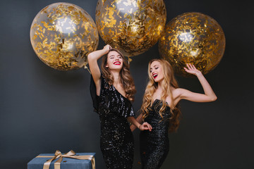 Naklejka premium Beautiful ladies dancing with hands up in front of shining helium balloons and smiling. Indoor photo of refined brown-haired birthday girl chilling with friend and laughing.