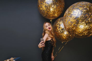 Naklejka premium Indoor portrait of stunning fair-haired girl making selfie at festive. Spectacular woman with party balloons having fun and taking photo of herself on black background.