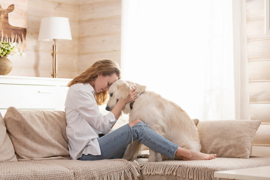 Young pretty woman in casual clothes hugging her beloved big white dog sitting on the sofa in the living room of her cozy country house. Animal communication concept