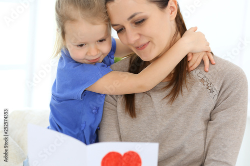 Mother's day concept. Child daughter congratulates mom and gives her postcard with red heart shape. Mum and girl happy smiling and hugging. Family fun and holiday
