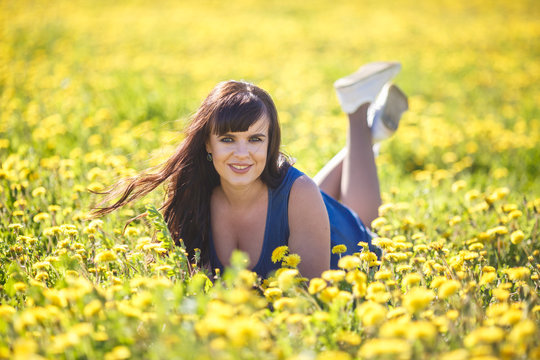 young happy beautiful big size model girl in blue dress lies resting on blooming dandelions field in summer.