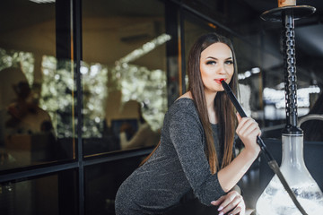 Young beautiful girl sits and smokes hookah on the summer terrace of a modern cafe.