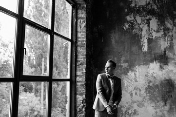 Morning of the groom. Waiting for the bride. The groom smiles in a gray suit and a bow tie. He repairs his jacket and looks out the window. Room in loft style.  Black and white photo