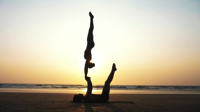 Silhouette of fit sporty couple practicing acrobatic yoga with partner together on the sandy beach. Female acrobat doing handstand on the hands of his lying male partner. 