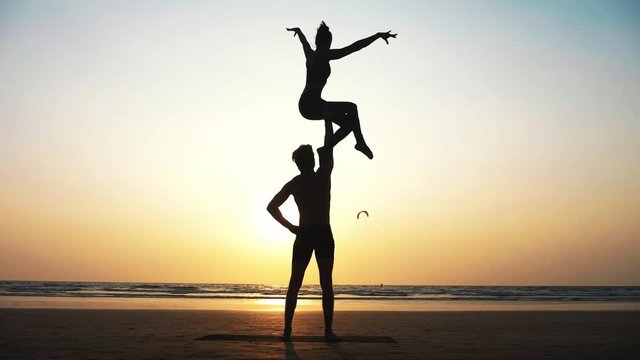 Silhouette of fit sporty couple practicing acrobatic yoga with partner together on the beach. Male standing and holding his female partner over his head. Mix race couple doing acrobatic exercise. 