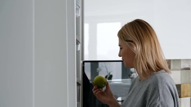 Cute young blonde girl takes a green apple from the fridge to make a snack. Concept of proper nutrition and healthy snack