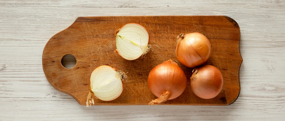 Unpeeled raw organic golden onions on rustic wooden board over white wooden background, overhead view. Flat lay, top view, from above.