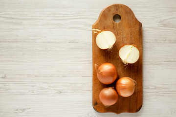 Unpeeled raw organic yellow onions on rustic wooden board over white wooden surface, top view. Flat lay, overhead, from above. Copy space.