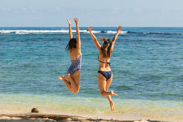 Two attractive joyful girls in blue bikini and long hair jumping on the beach with hands outstretched expressing happiness in vacation.