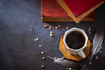 Black coffee in white cup and old books with feather and dried flower petals placed on the marble table and morning sunlight.
