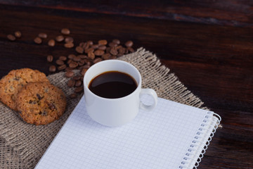 White cup of coffee with coffee grains near and biscuits and notepad in a cage for records on linen fabric on the dark vintage background