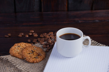 White cup of coffee with coffee grains near and biscuits and notepad in a cage for records on linen fabric on the dark vintage background
