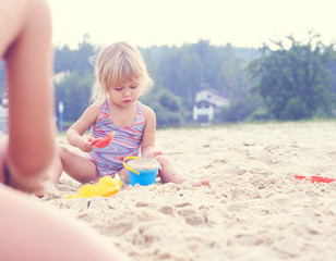 Little blonde baby girl playing with sand on the beach using plastic toys.
