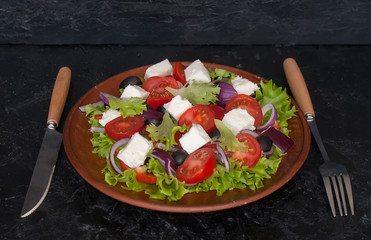 Traditional Greek salad with vegetables and feta cheese in a clay bowl on a black slate with Cutlery.