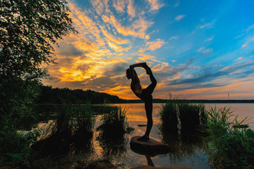 Blonde woman doing bow pulling pose outdoors on a stone in the lake. Yoga nature concept.