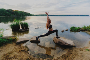Blonde woman doing anjaneyasana lunge outdoors on a rocks in lake. Yoga nature concept.