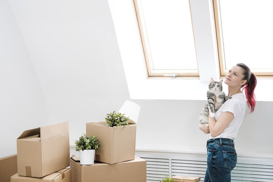 Young beautiful girl with colored hair in a white T-shirt and jeans, holding a pet cat and looking out the window against the background of cardboard boxes and things.