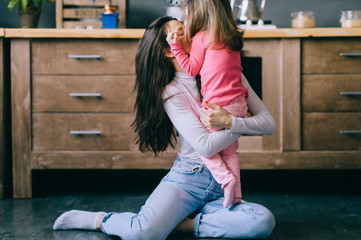Adorable young woman playing with her little funny daughter in kitchen. Portrait of pretty mother hugging, carrying and watching her litle child. Happy female family indoor lifestyle portrait.