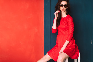 Beautiful fashion girl with long hair, spanish appearance in sunglasses and red elegant dress posing on blue red wall in studio. Amazing babe in suglasses with black hair. Hairstyle advertsing.